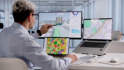 A male office worker sits at a white desk with multiple screens displaying GIS mapping data, pointing a stylus at a large monitor showing a 3D urban model and data charts.